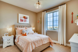 A peach colored girls bedroom with comfortable bed featuring many pillows and a large windows.