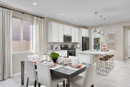 A dining room featuring ahite and orange accents on the table, leading into a brand new kitchen with white cupboards and an island.