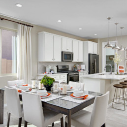 A dining room featuring ahite and orange accents on the table, leading into a brand new kitchen with white cupboards and an island.