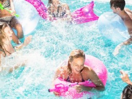 A group of young adults on flotation devices, laughing and spraying each other with water guns in a splashing pool.