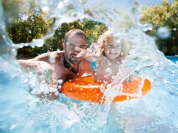 A young child with an orange inner tube and their father playing and splashing in a pool.
